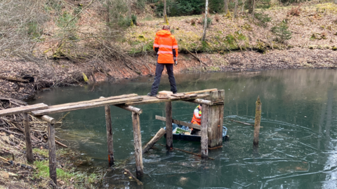 Arbeitseinsatz mit Freiwilligendienstleistenden in der Teichpflege  Auf dem Foto sehen Sie einen Freiwilligendienstleistenden auf einem Steg. Ein anderer Sitz auf einem Boot auf einem Teich.