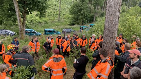 Eine Gruppe Förster in Signalkleidung steht im Wald und hört gespannt den Ausführungen des Dozenten zu. 
