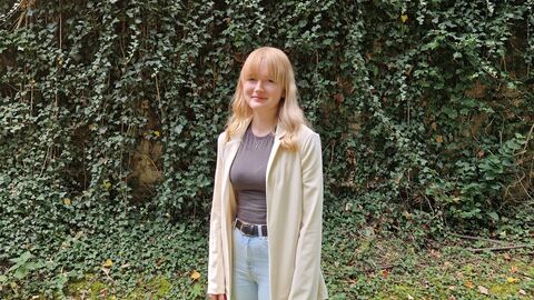 Eine junge Frau steht in einem Garten vor einer bewachsenen Mauer und lächelt. 