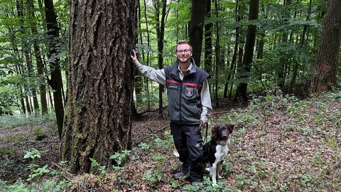 Ein junger Förster steht mit seinem Jagdhund an einer starken Lärche, berührt sie mit seiner Hand und lächelt in die Kamera. 