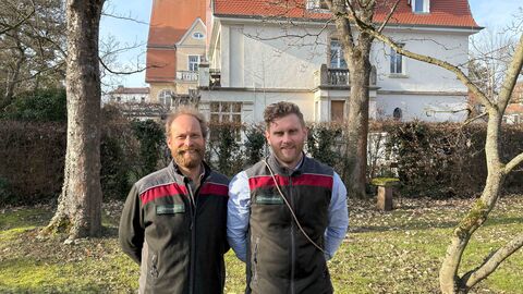 Zwei Männer stehen in einem Garten vor eine Villa. Sie tragen Forstdienstkleidung und lächeln den Betrachter an. 