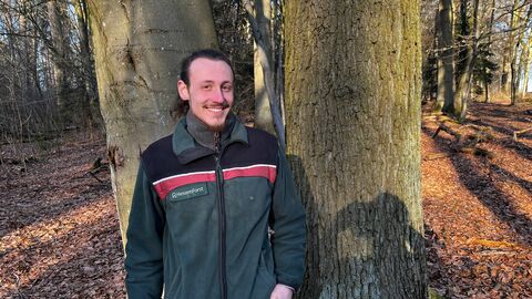 Ein Mann in Forstdienstkleidung steht vor zwei Baumstämmen im herbsltichen Wald und lächelt den Betrachter an.
