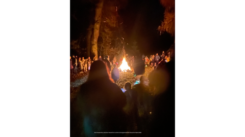 Eine Menschengruppe steht im dunkeln um ein Lagerfeuer.