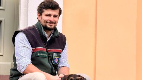 Ein junger Man sitzt auf einer kleinen Treppe vor einer Tür mit seinem Dackel und schaut den Betrachter an. 