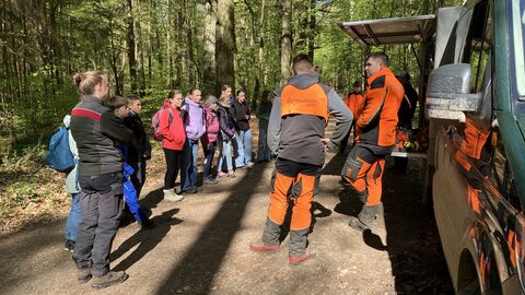 Mehrere MEnschen stehen in Schutzkleidung im Wald. Dazwischen kinder und ein Forstfarzeug. 