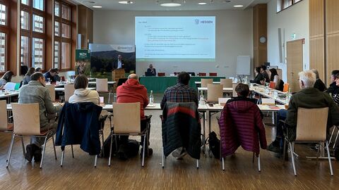 Eine Gruppe von Personen sitzt in einem Besprechungssaal. Auf einer LEinwand ist eine Vortragsfolie zu sehen. 