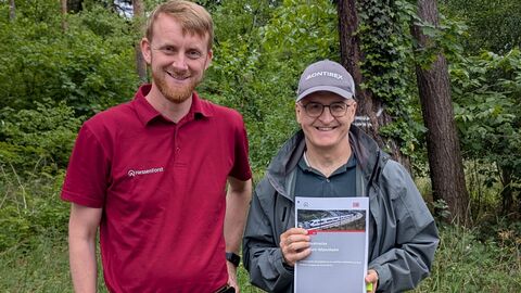 Zwei Männer stehen vor einem Waldrand und präsentieren lächelnd ein Schriftsück.