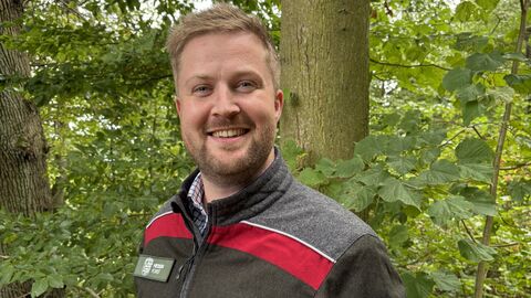 Ein junger mann in Forstdienstkleidung steht vor einem Waldhintergrund aus grünen Blättern und Bäumen